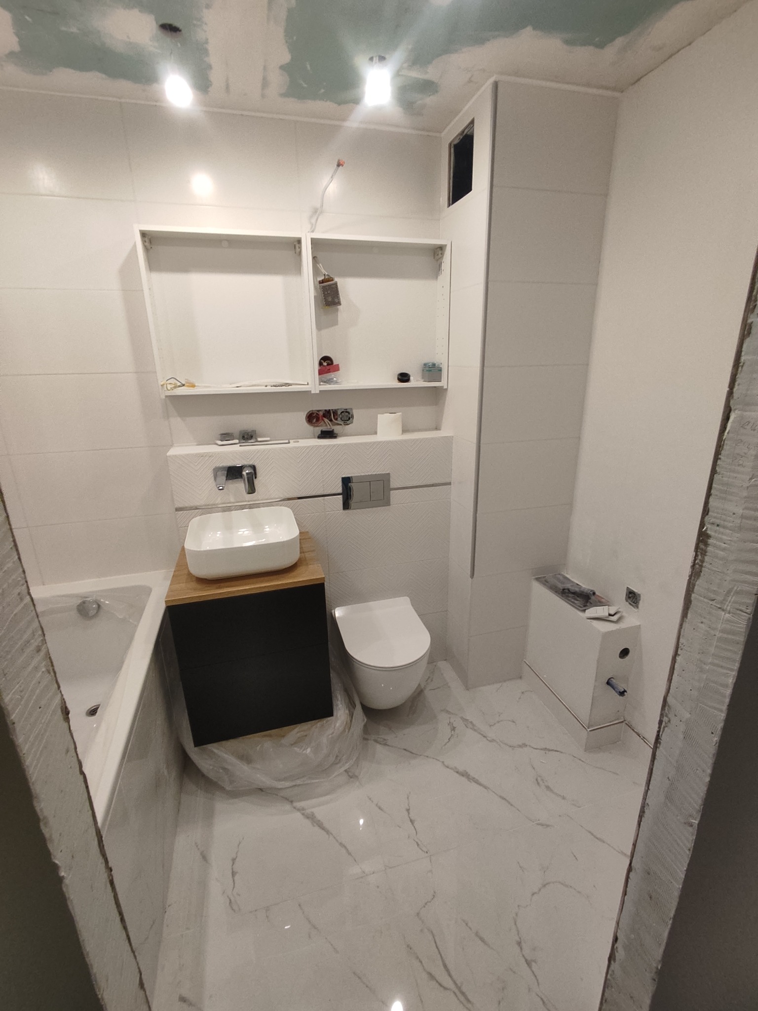 Wnętrze łazienki w trakcie remontu: białe płytki na ścianach, marmurowa podłoga, umywalka nablatowa z drewnianym blatem, podwieszana toaleta, otwarte półki i częściowo wykończony sufit.