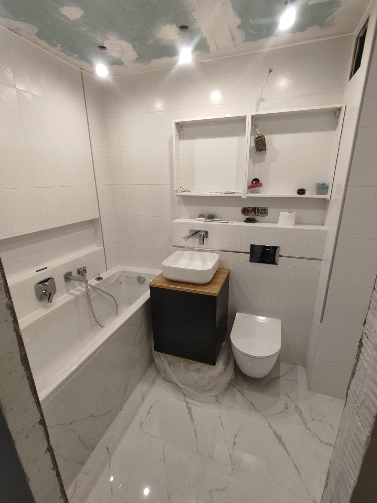 Biała łazienka w trakcie wykańczania: położone płytki, zamontowana wanna, umywalka na szafce i toaleta, sufit wymaga jeszcze prac wykończeniowych.