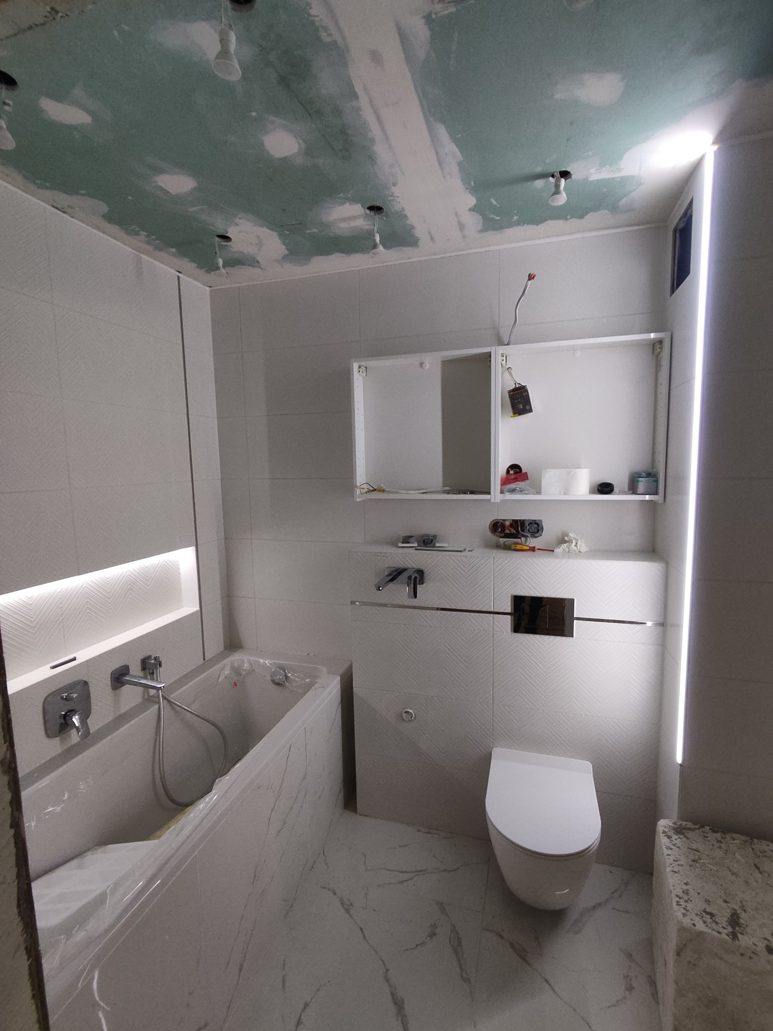 Wnętrze łazienki w trakcie remontu, widoczne białe płytki z delikatnym wzorem, częściowo zakryta folią wanna, podwieszana toaleta, odkryta sufitowa instalacja elektryczna i niezakończone prace...