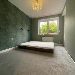 Pomalowane na zielono wnętrze sypialni z dekoracyjną fakturą na ścianie, widoczne dwa czarne reflektory ścienne, materac na podłodze, szary dywan i okno z widokiem na drzewa.