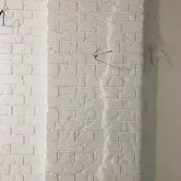 Fragment ściany z białej cegły dekoracyjnej, częściowo pomalowanej na jaśniejszy odcień bieli, z widocznymi przewodami elektrycznymi i śladami po pracach wykończeniowych na sąsiedniej ścianie.