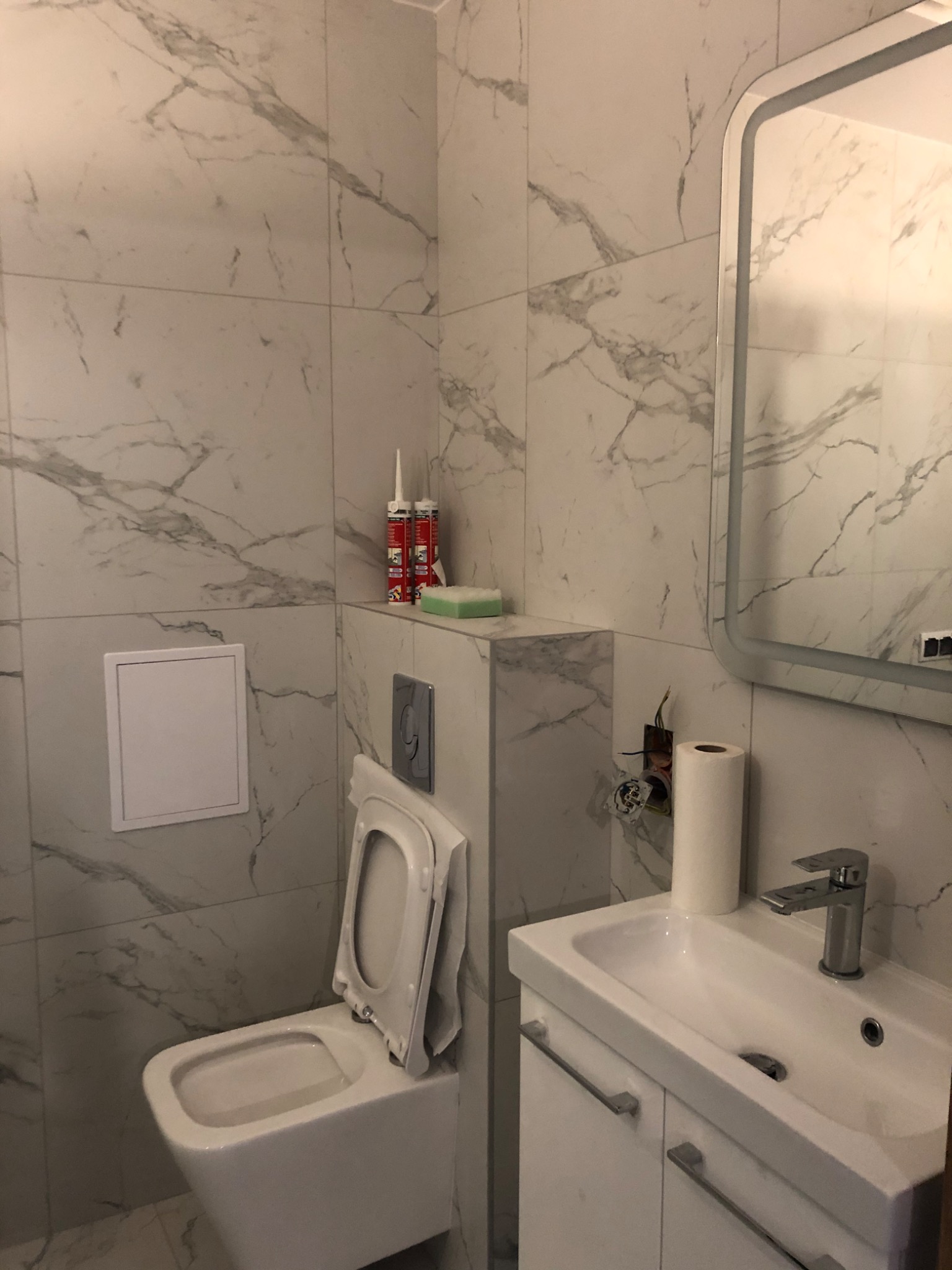 Wykończona łazienka z białymi płytkami imitującymi marmur, białą toaletą z podniesioną deską, umywalką z szafką i podświetlanym lustrem. Widoczne akcesoria montażowe i przewody elektryczne przed...