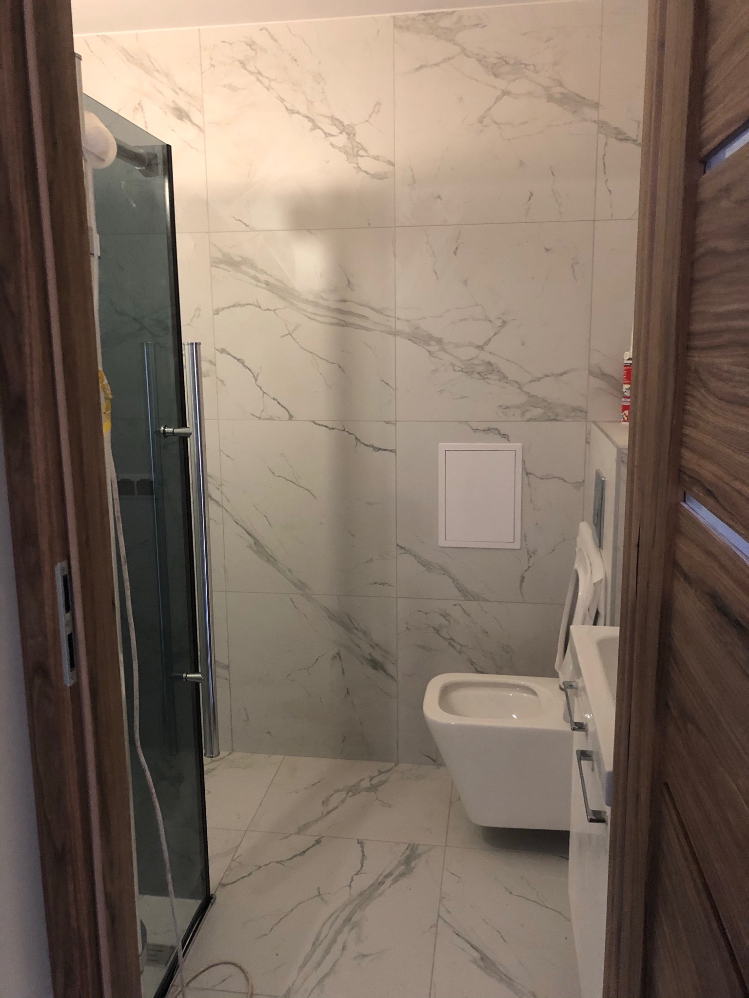 Wykończona łazienka z kabiną prysznicową i podwieszaną toaletą, białe płytki imitujące marmur na ścianach i podłodze, widok zza drewnianych drzwi.