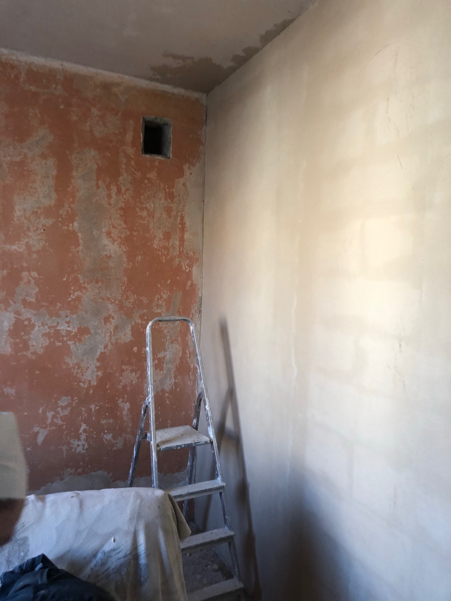 Pokój w trakcie remontu: ściana z fragmentami starej farby, obok ściana po nałożeniu wstępnej warstwy gładzi, drabina malarska w rogu.