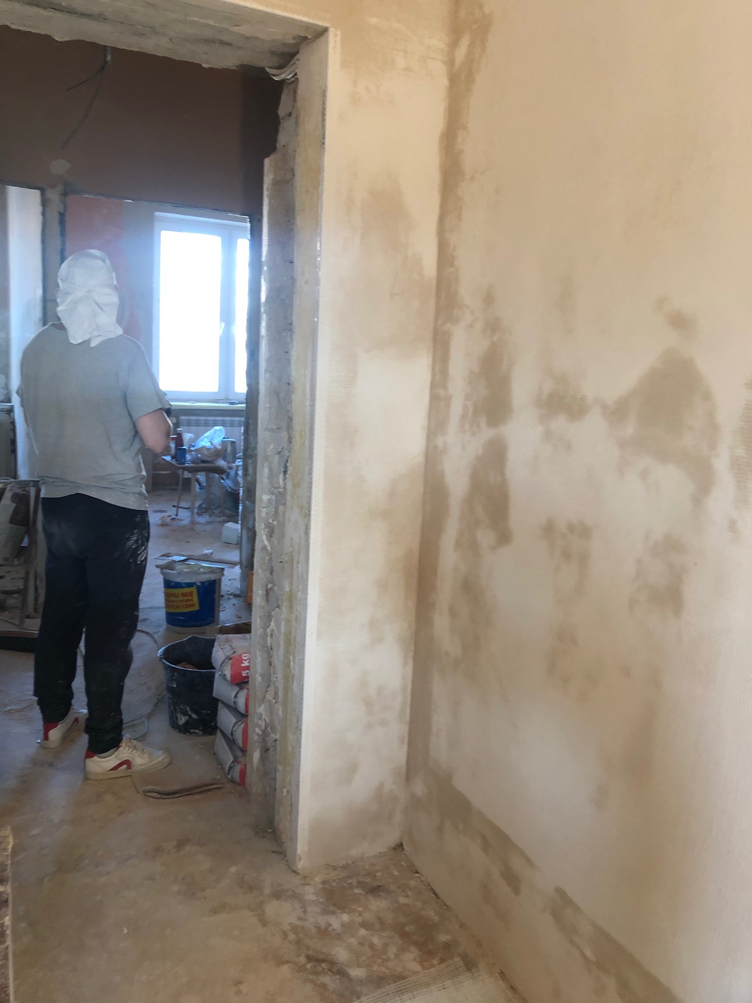 Wnętrze w trakcie prac wykończeniowych: widok na ścianę z nałożoną warstwą wyrównującą, pracownik w tle, worki z materiałem budowlanym i narzędzia.