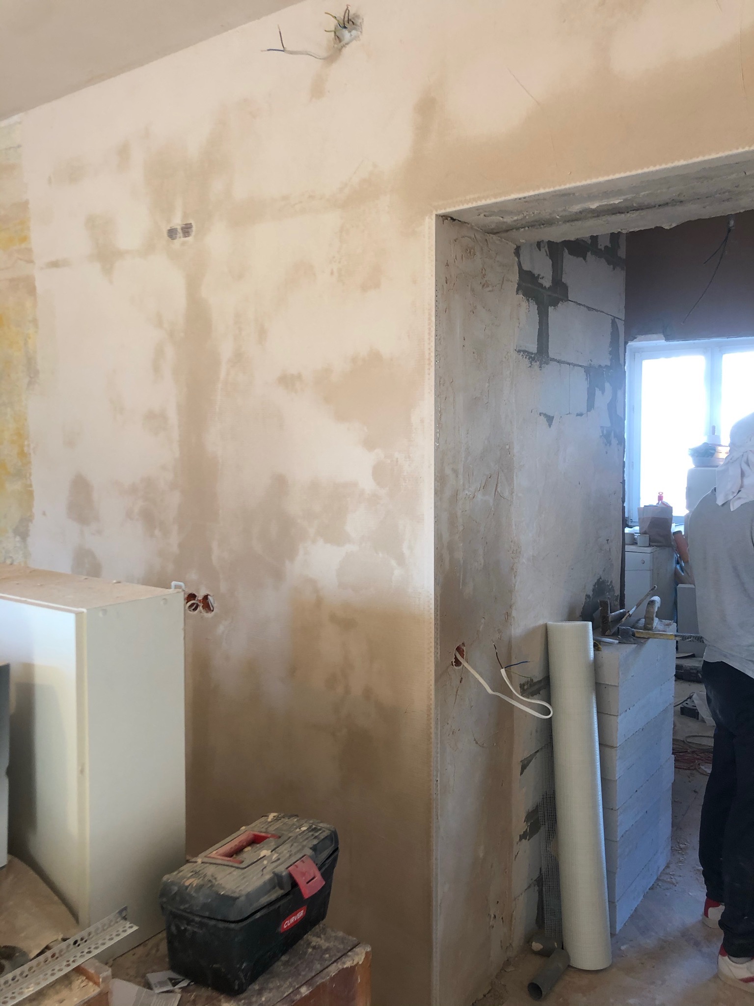 Wnętrze w trakcie prac wykończeniowych: częściowo nałożona warstwa wyrównująca na ścianie, widoczne puszki elektryczne, narzędzia i materiały budowlane, fragment murowanej ściany w tle.