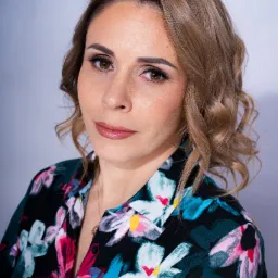 Portret kobiety z brązowymi włosami i w kwiecistej bluzce, subtelny makijaż, neutralne tło.