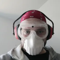 Portret pracownika remontowego w okularach ochronnych, masce przeciwpyłowej i słuchawkach, z czerwoną bandaną na głowie, patrzącego w dół na tle białej ściany.