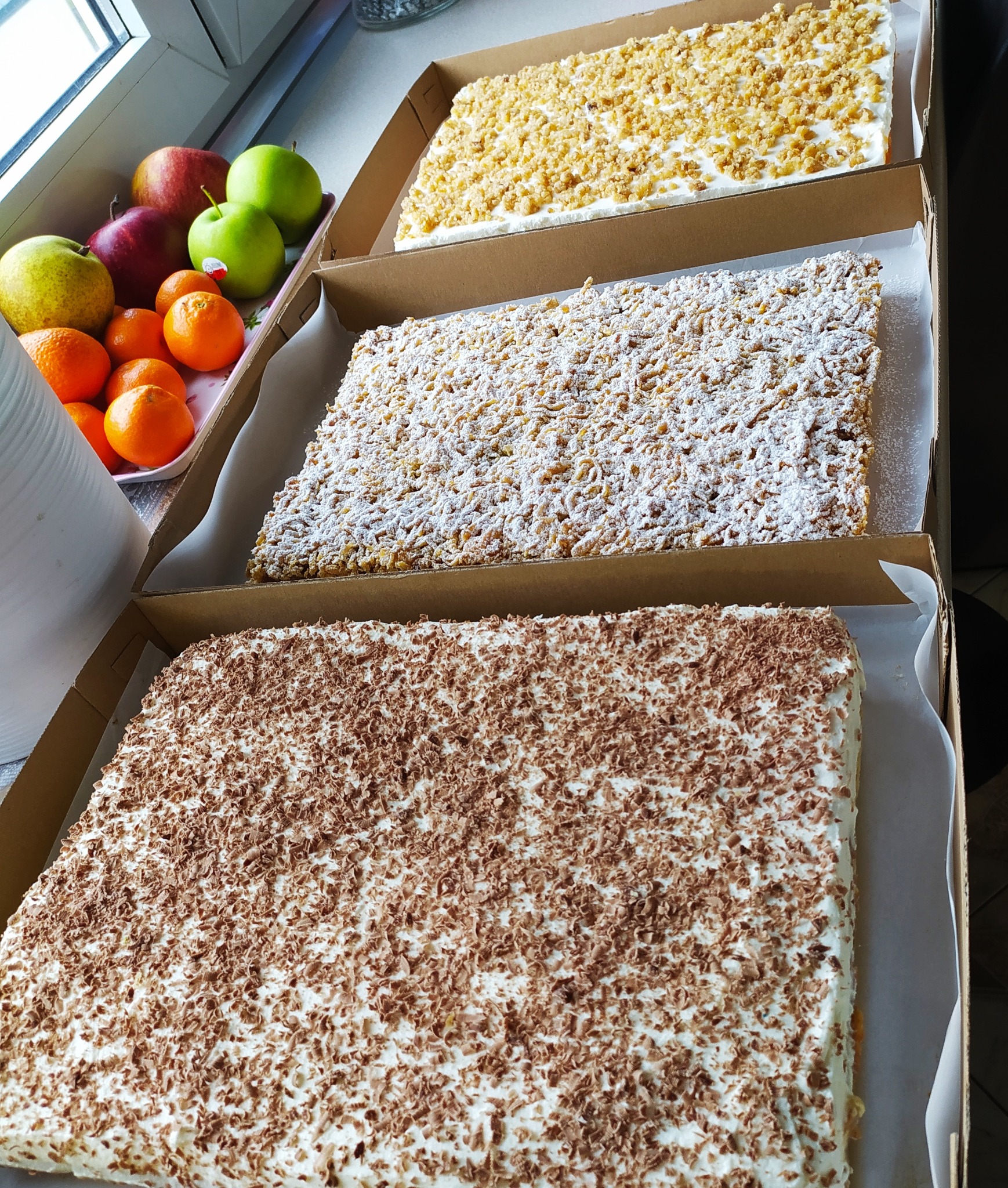 Trzy prostokątne torty w kartonowych pudełkach: jeden z posypką z kruszonki, drugi z kruszonką posypaną cukrem pudrem, trzeci z posypką z wiórków czekoladowych; obok taca z jabłkami, gruszkami...