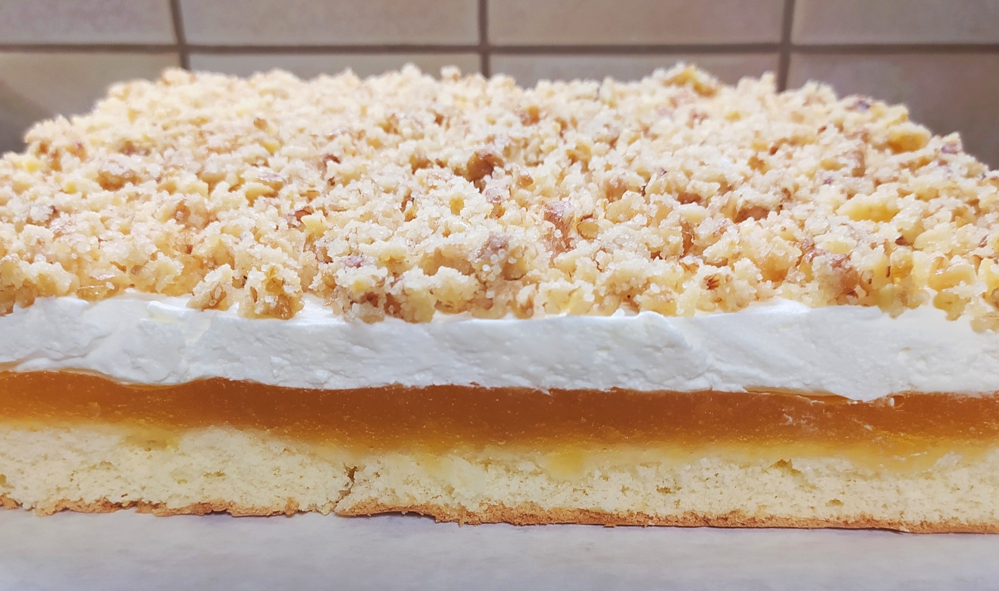 Przekrój ciasta z galaretką brzoskwiniową, bitą śmietaną i kruszonką orzechową na jasnym biszkopcie, sfotografowane z bliska.