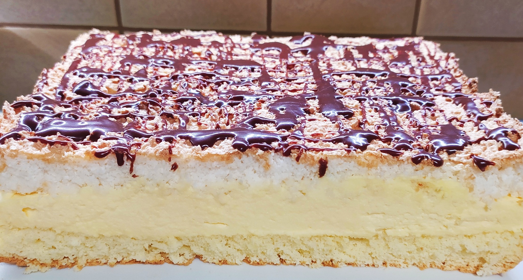 Przekrój prostokątnego ciasta z kremem, warstwą bezy i polewą czekoladową.