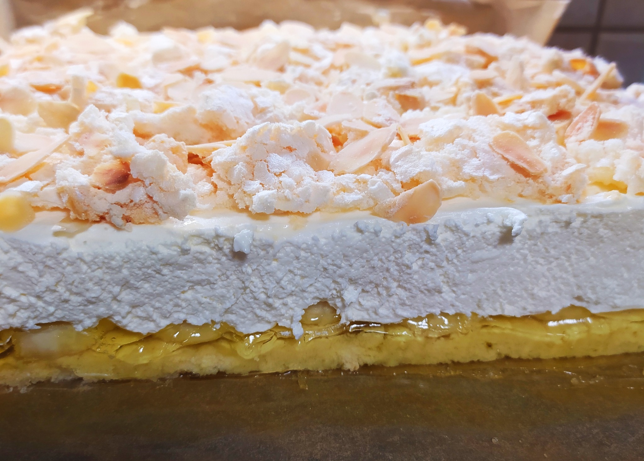 Przekrój tortu z kremem, warstwą galaretki i posypką z bezy i płatków migdałów, widoczny na blacie pokrytym papierem do pieczenia.