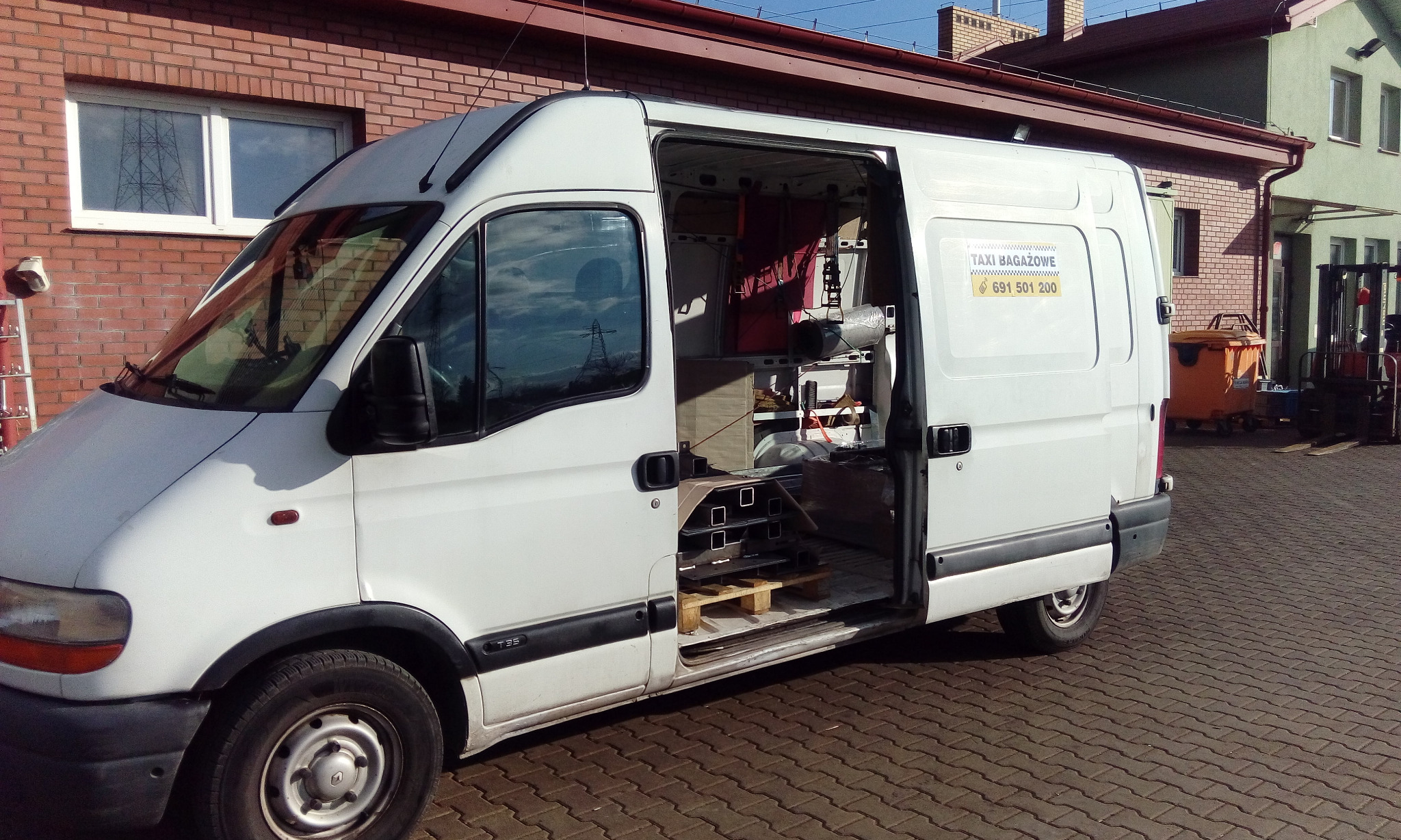 Biały bus z otwartymi drzwiami bocznymi, wewnątrz widoczne metalowe profile na palecie, na boku naklejka 'Taxi Bagażowe' z numerem telefonu, w tle budynki i wózek widłowy.
