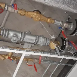 Widok z dołu na zamontowane rury hydrauliczne i grzewcze pod sufitem, widoczne zawory z czerwonymi kurkami, izolacja na rurach i elementy mocujące do sufitu.