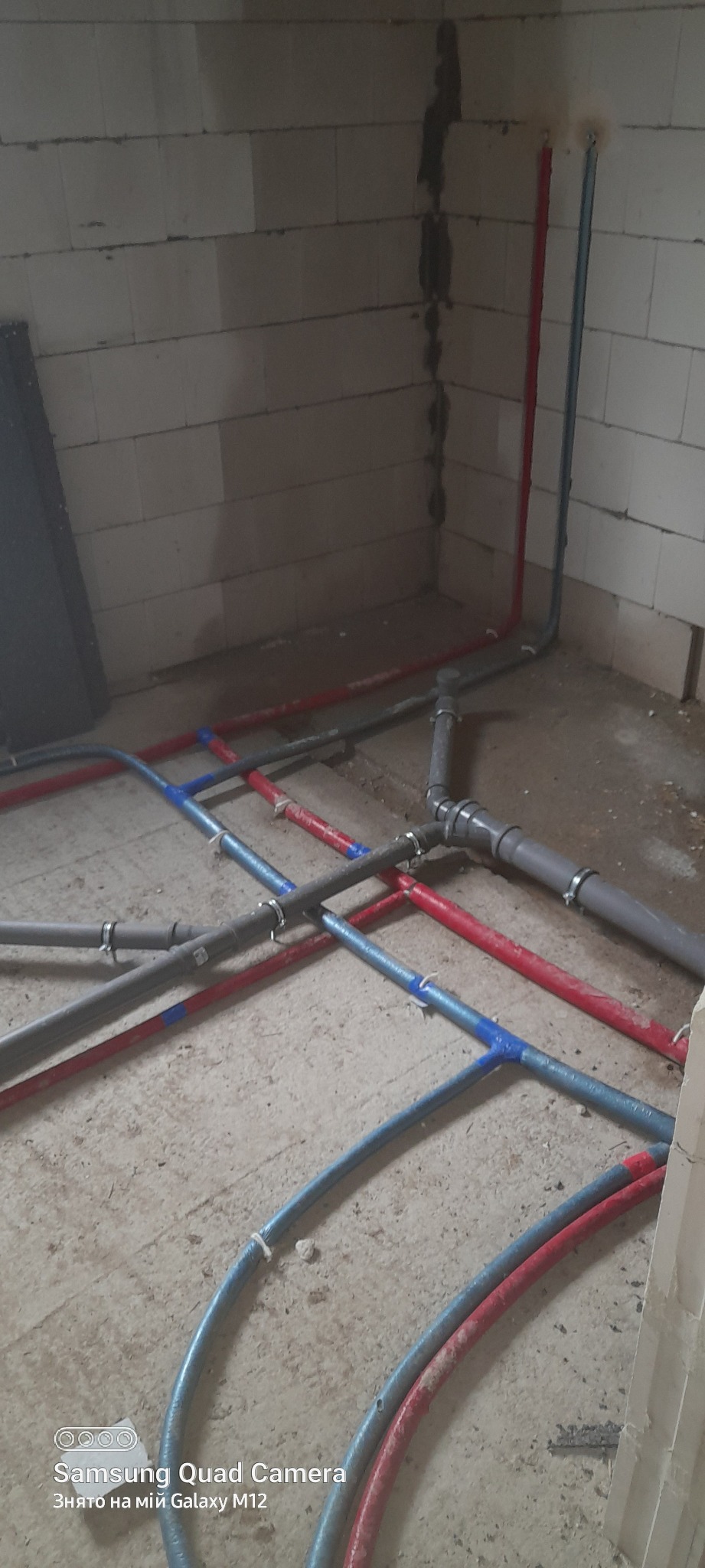 Instalacja wodna i kanalizacyjna w surowym stanie, widoczne rury PEX w kolorze czerwonym i niebieskim oraz szare rury kanalizacyjne, połączone złączkami, zamocowane do podłoża za pomocą uchwytów.