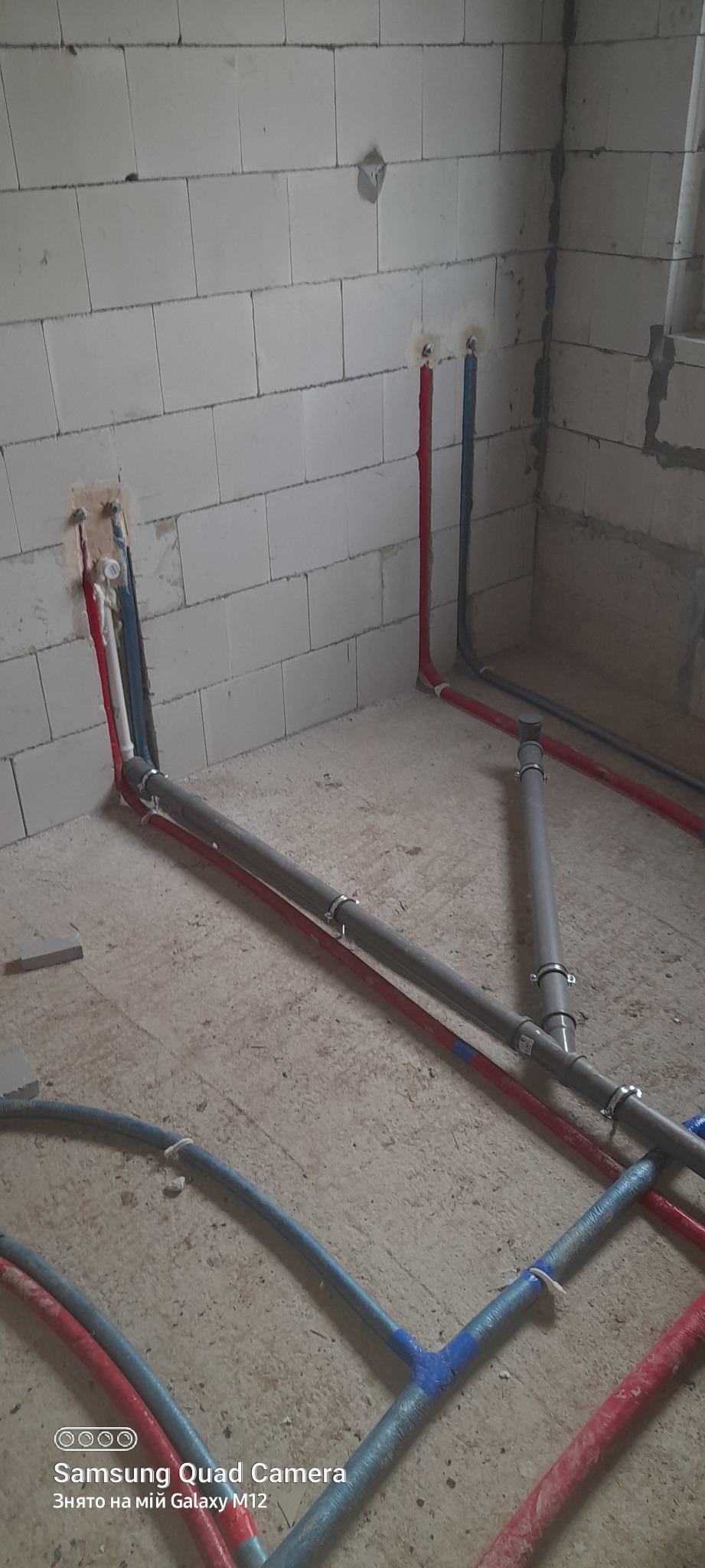 Wewnętrzna instalacja wodno-kanalizacyjna w surowym stanie, widoczne rury w kolorach czerwonym i niebieskim prowadzone po podłodze i w ścianach z betonu komórkowego.