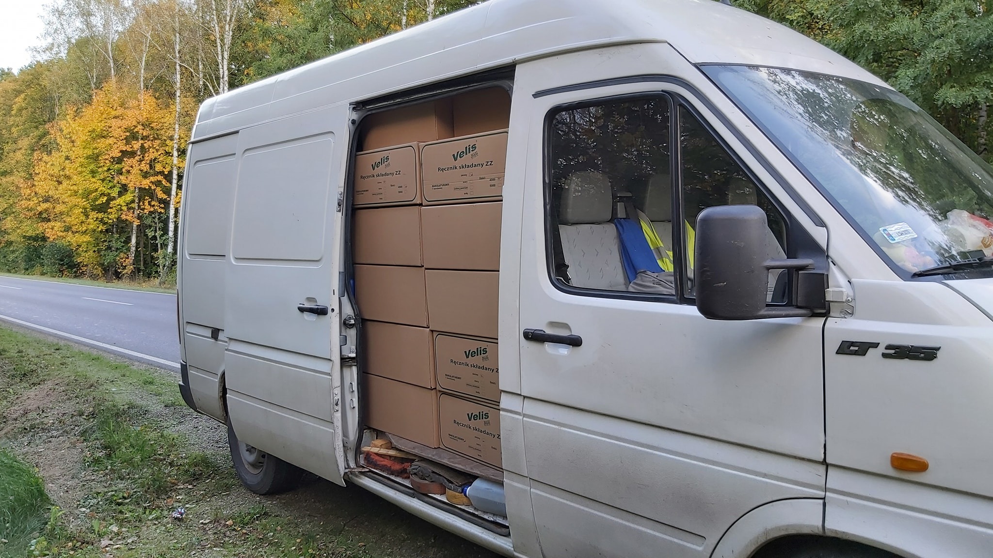 Biały bus z otwartymi drzwiami bocznymi, wypełniony kartonami z napisem 'Velis Ręcznik składany ZZ', zaparkowany na poboczu drogi w pobliżu lasu z jesiennymi drzewami w tle.