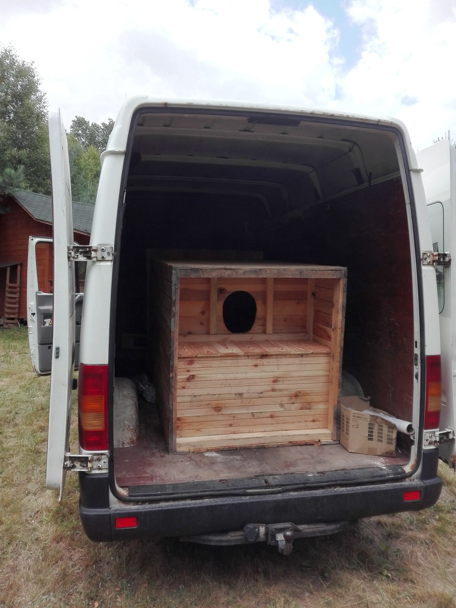 Drewniana budka z okrągłym otworem umieszczona w przestrzeni ładunkowej białego busa z otwartymi drzwiami, widoczna kartonowa paczka obok budki.