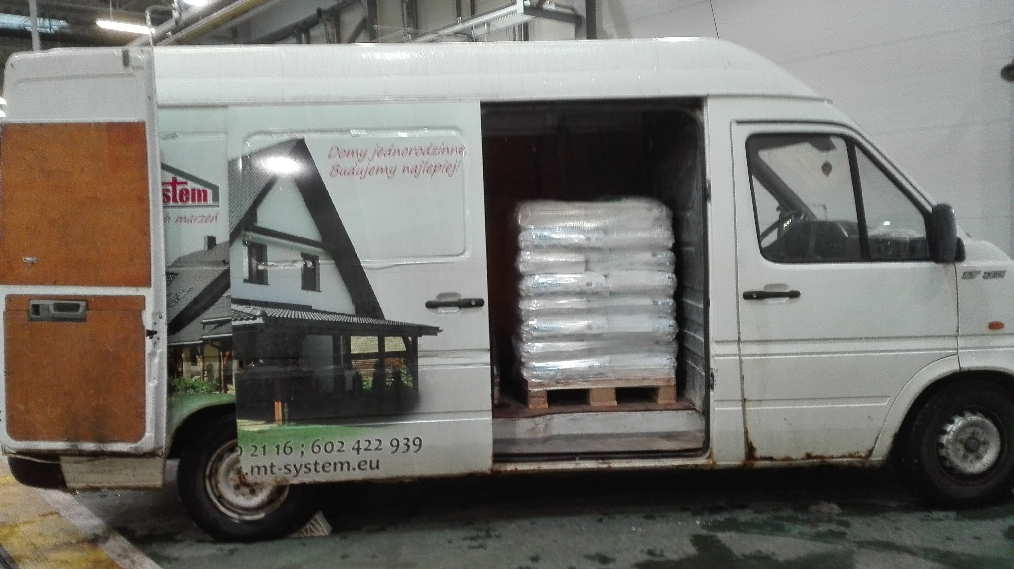 Biały bus z otwartymi drzwiami bocznymi i tylnymi, załadowany paletą z workami, na karoserii reklama firmy budowlanej z numerem telefonu.