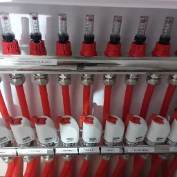 Rozdzielacz instalacji podłogowej z czerwonymi rurami i etykietami pomieszczeń: łazienka, pokój córki, korytarz, sypialnia, biuro, salon i kuchnia. Widoczne przepływomierze z czerwonymi elementami...