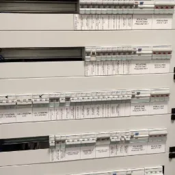 Rozbudowana tablica rozdzielcza z licznymi wyłącznikami, bezpiecznikami i stycznikami, oznaczona etykietami opisującymi obwody i funkcje, w szarej obudowie.