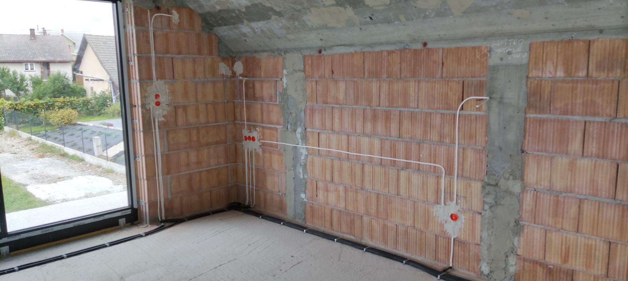 Wewnętrzne okablowanie elektryczne w surowym stanie, białe przewody w peszlach na ścianach z czerwonej cegły i podłodze, widoczne puszki instalacyjne.