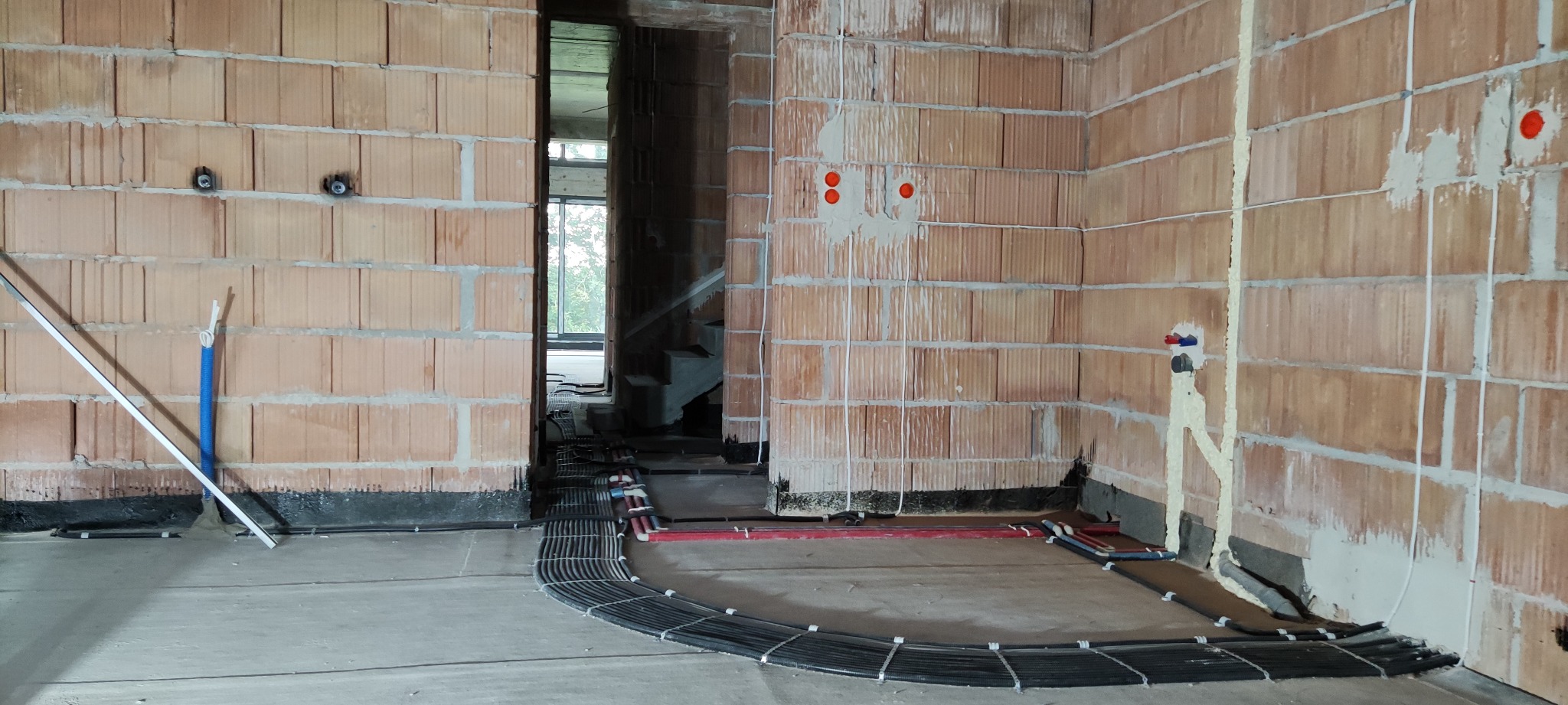 Surowy stan wnętrza z widocznymi przewodami elektrycznymi ułożonymi wzdłuż ściany z cegły ceramicznej, wystające kable i puszki instalacyjne, pianka montażowa wokół rur.
