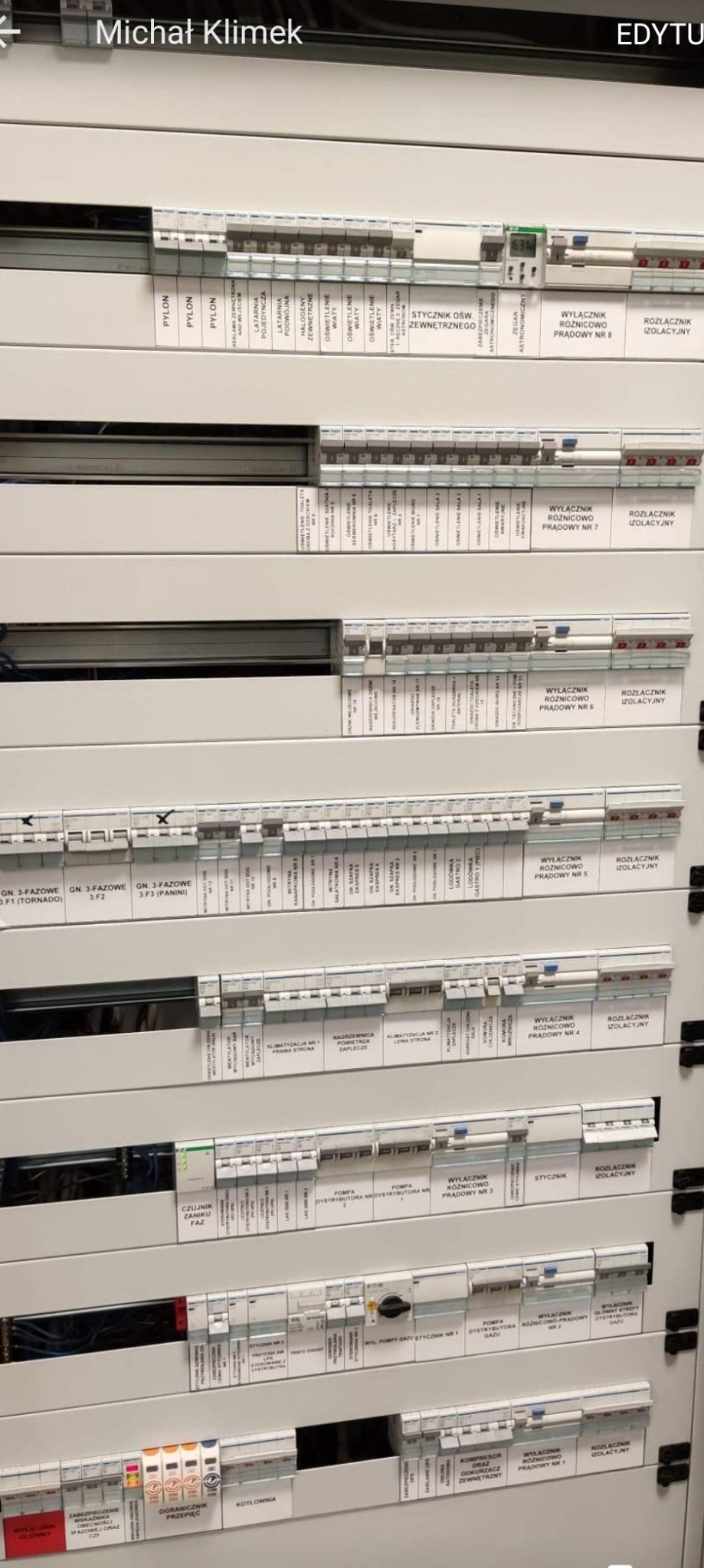 Rozbudowana tablica rozdzielcza z licznymi wyłącznikami, bezpiecznikami i stycznikami, oznaczona etykietami opisującymi obwody i funkcje, w szarej obudowie.