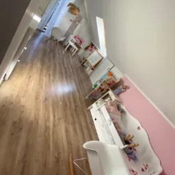 Widok na posprzątany pokój dziecięcy z drewnianą podłogą, białymi meblami, tablicą do rysowania i różową ścianą, oświetlony naturalnym światłem wpadającym przez okno.