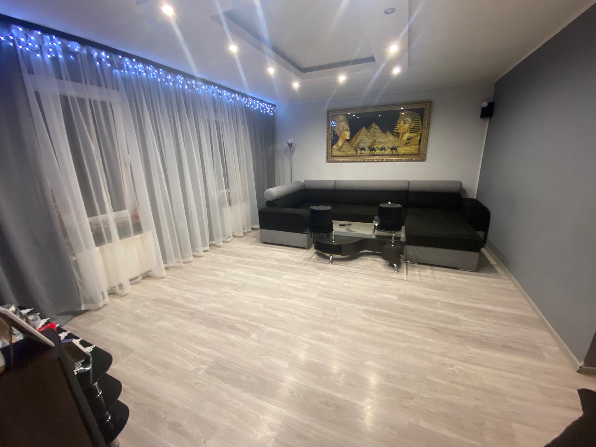 Nowoczesny salon z szarymi ścianami, narożną sofą, szklanym stolikiem kawowym i dekoracyjnymi lampkami LED nad oknem z firankami.