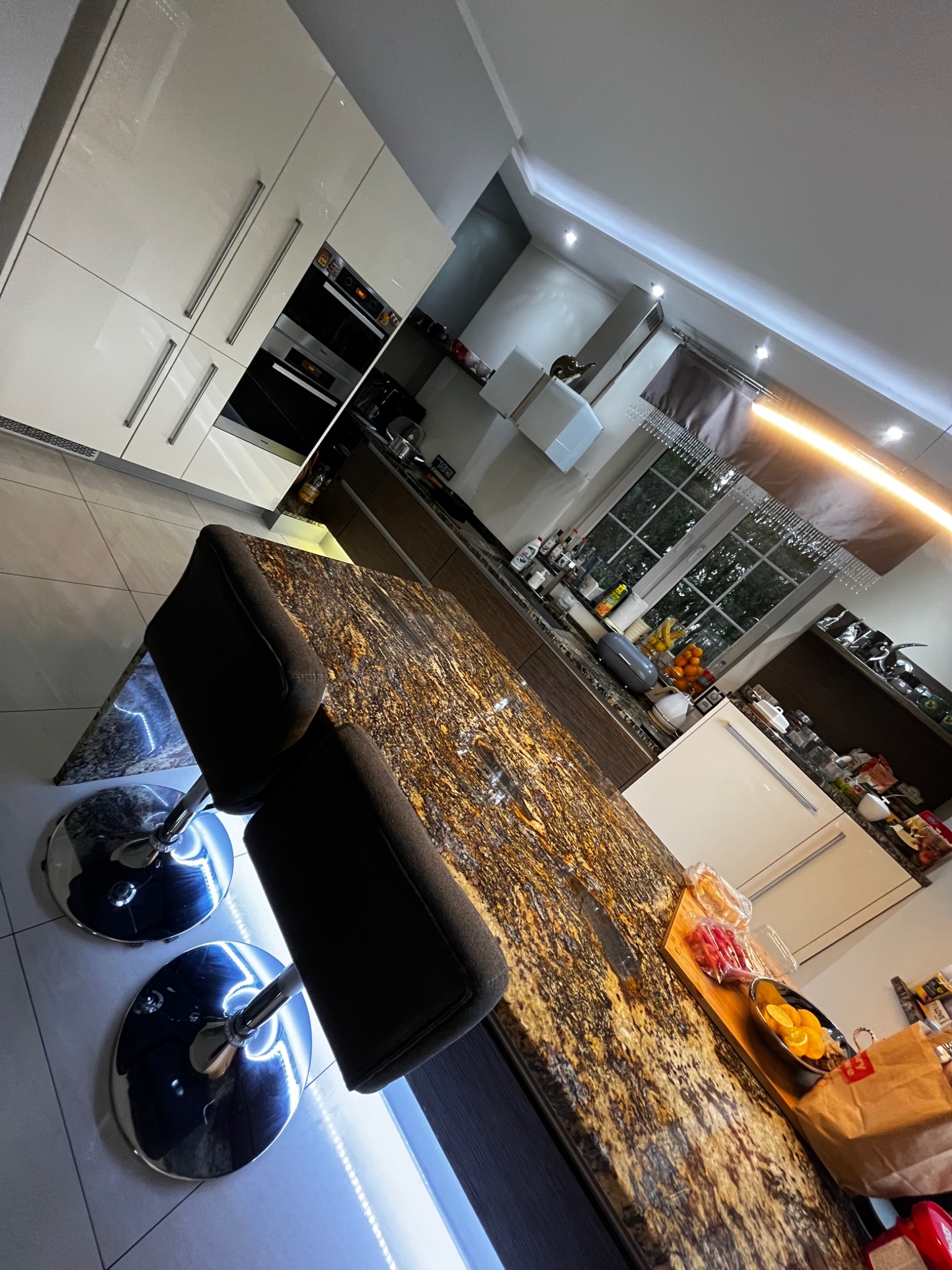 Nowoczesna, luksusowa kuchnia w Zabrzu z wyspą o marmurkowym blacie, widok z perspektywy baru z chromowanymi hokerami z podświetleniem LED, okno z firankami z koralików, jasne, lakierowane szafki.