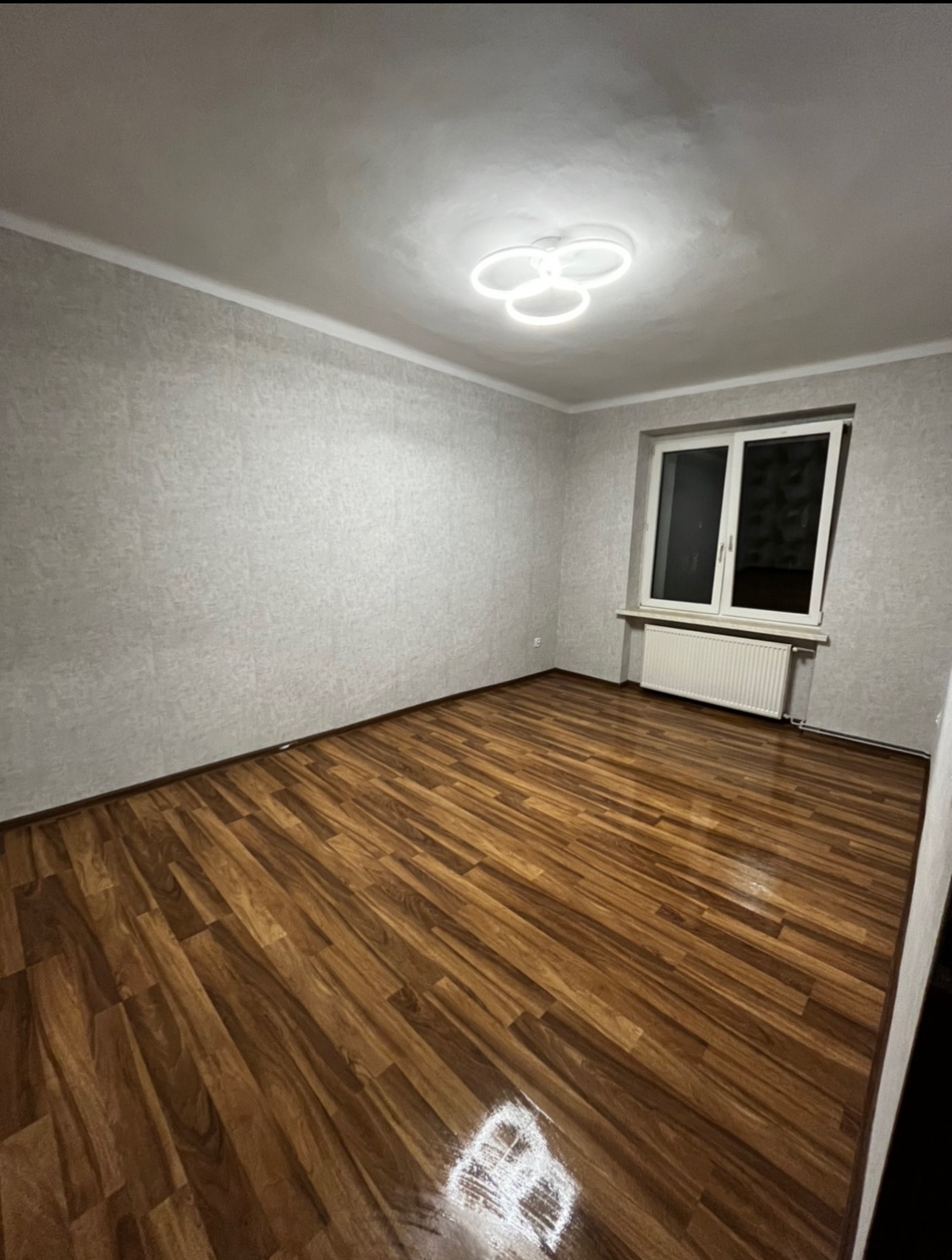 Puste pomieszczenie z brązowymi panelami podłogowymi, szarymi ścianami i nowoczesną lampą sufitową w kształcie trzech połączonych okręgów. Białe okno z grzejnikiem pod spodem.