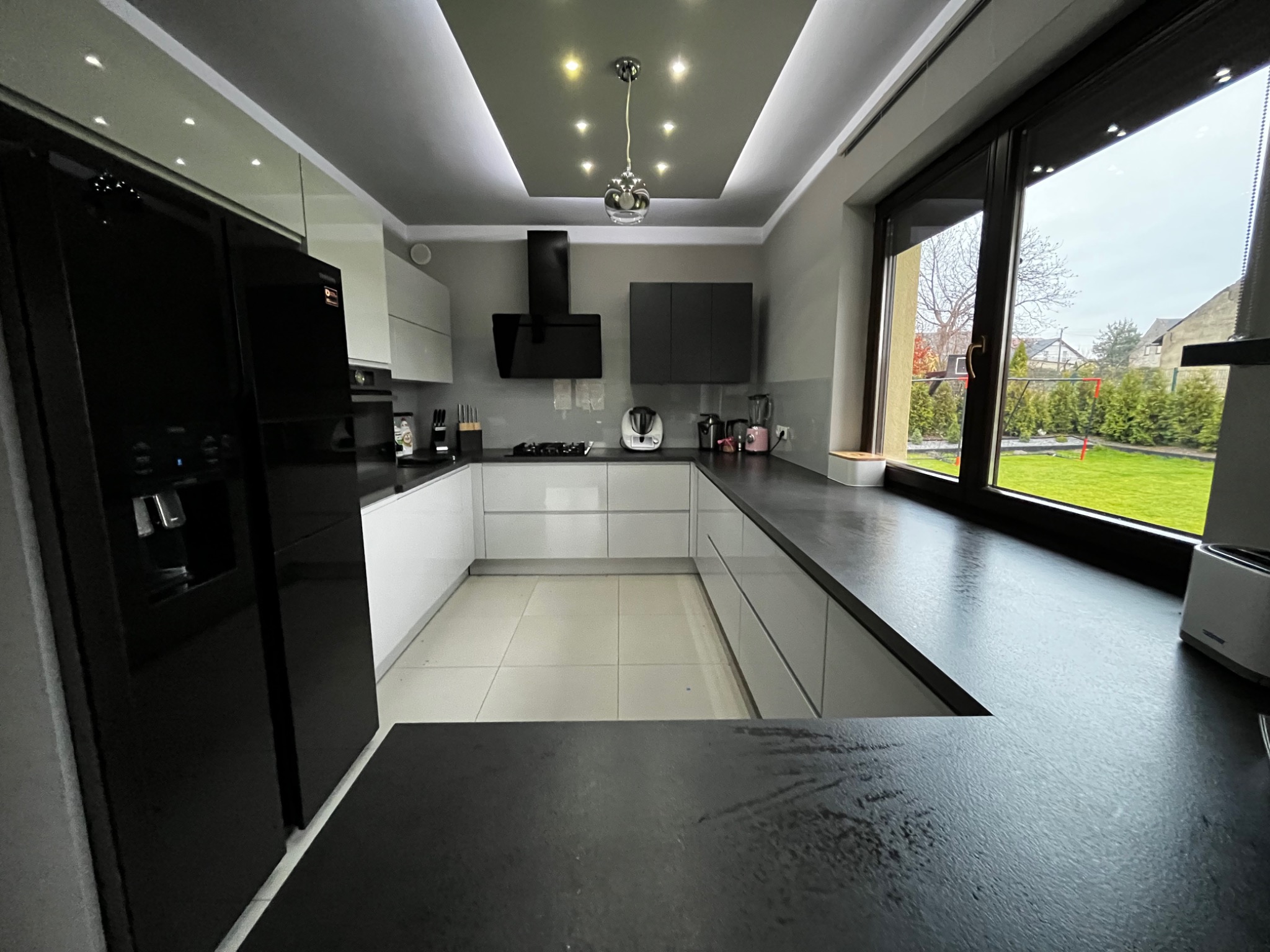 Nowoczesna, jasna kuchnia z czarnymi akcentami, widok z blatu na okno z widokiem na ogród, oświetlenie sufitowe z halogenami i wiszącą lampą.