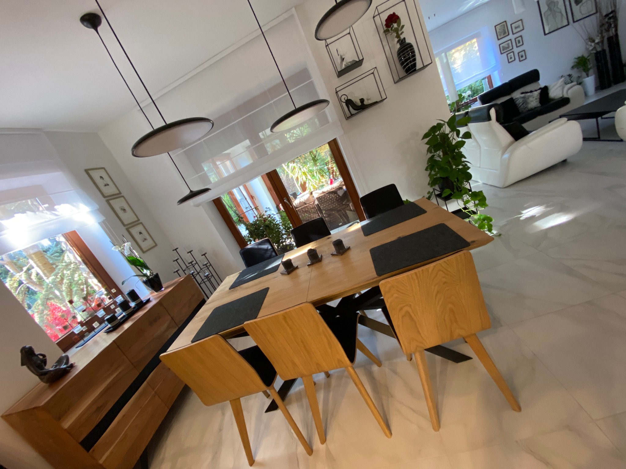 Jasne wnętrze jadalni z drewnianym stołem, krzesłami i komodą, uzupełnione czarnymi podkładkami, lampami wiszącymi i dekoracjami ściennymi, widoczne z perspektywy pod kątem.