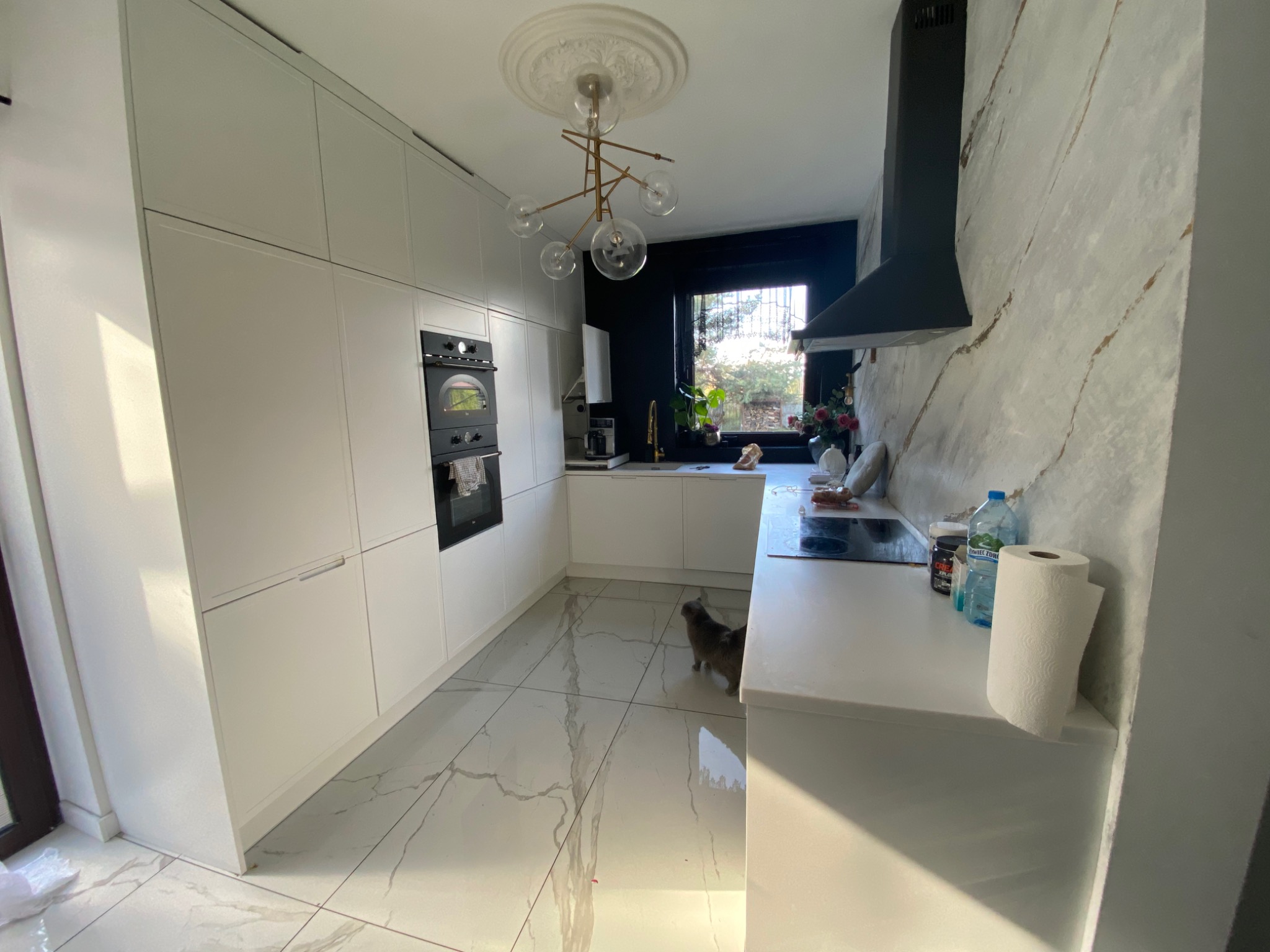 Nowoczesna, jasna kuchnia z białymi szafkami, marmurową podłogą i oryginalną lampą sufitową; kot w tle, butelki z wodą i ręcznik papierowy na blacie.