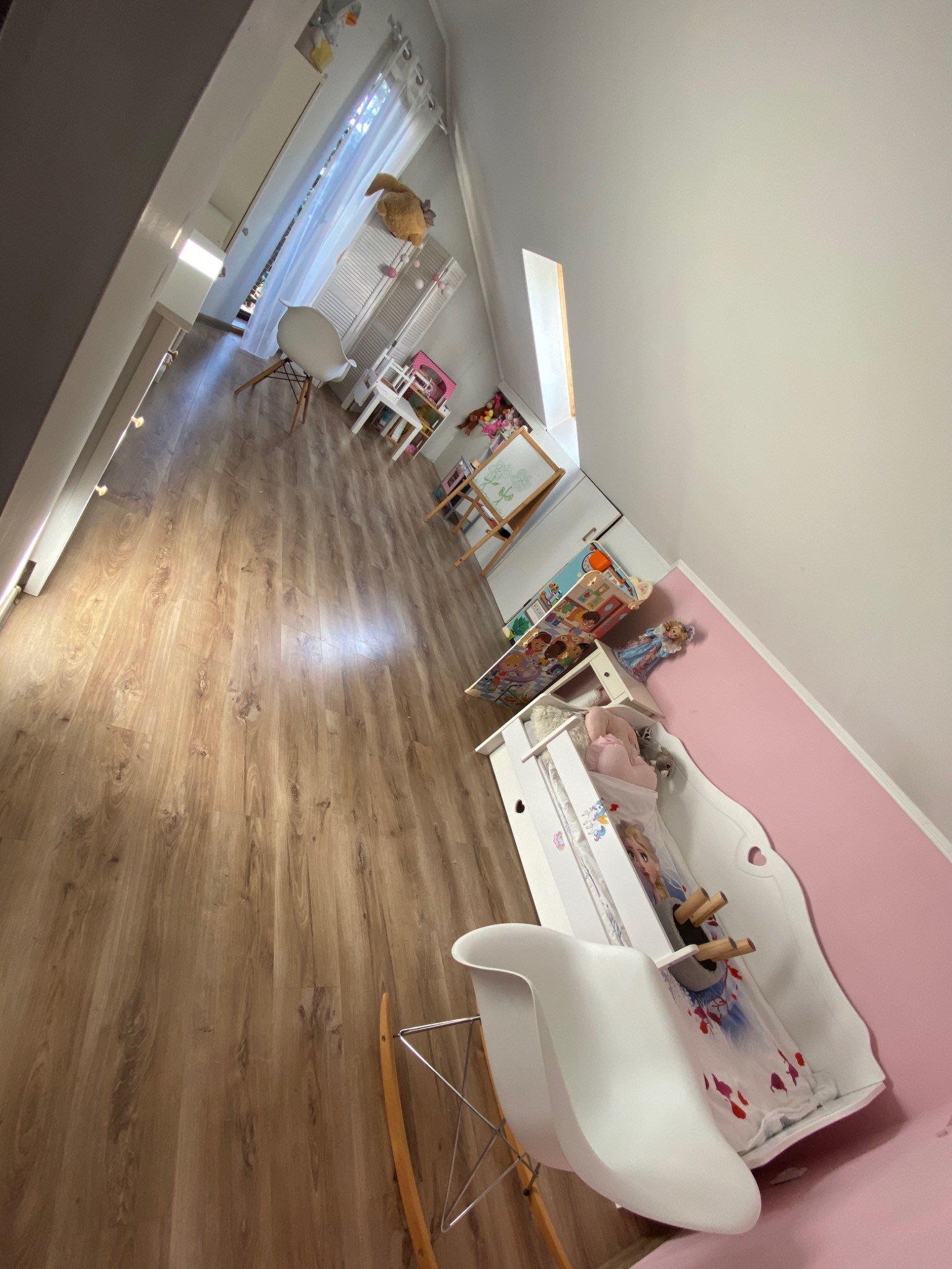 Widok na posprzątany pokój dziecięcy z drewnianą podłogą, białymi meblami, tablicą do rysowania i różową ścianą, oświetlony naturalnym światłem wpadającym przez okno.
