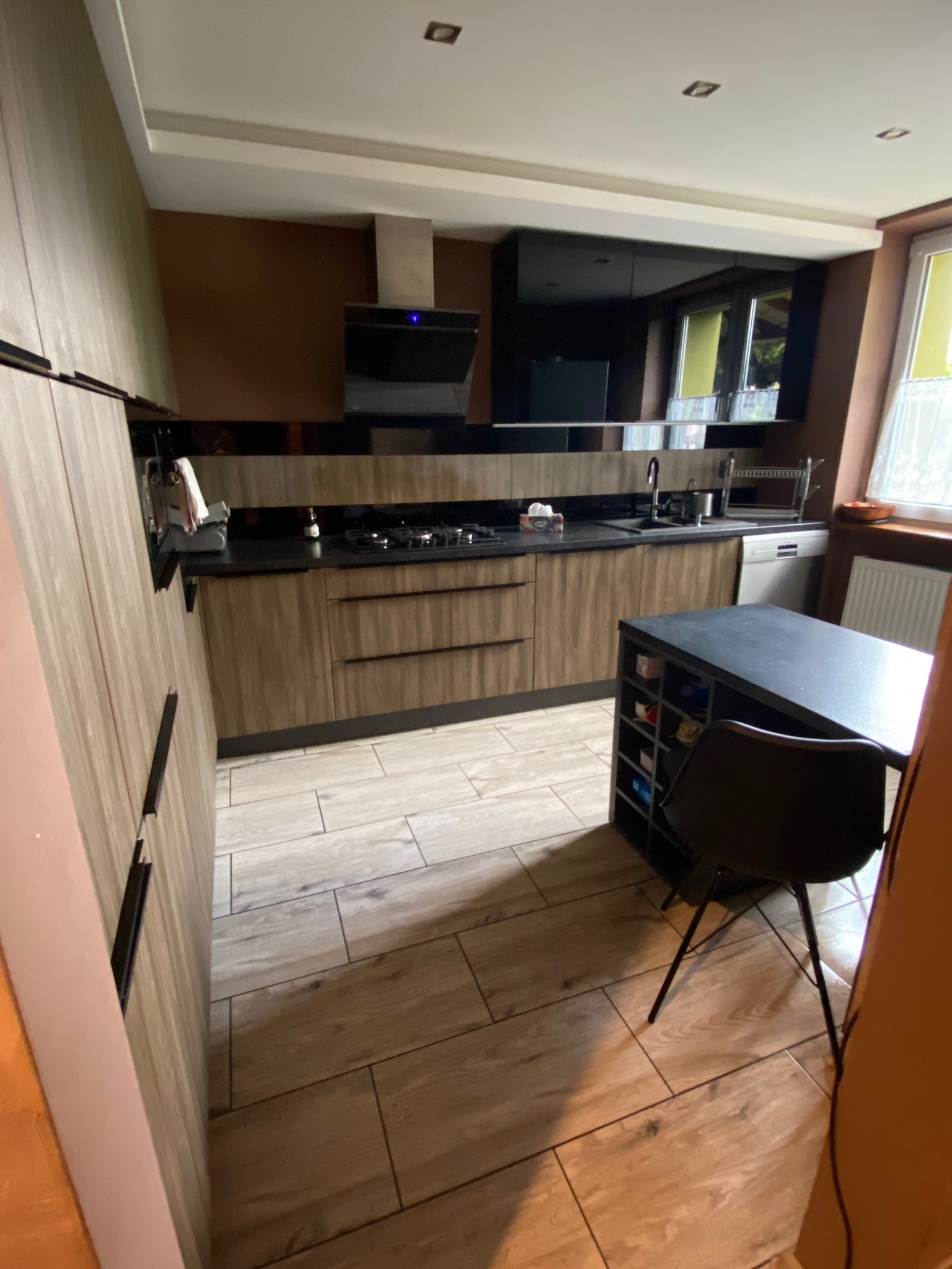 Nowoczesna kuchnia w odcieniach brązu i czerni, z drewnopodobnymi frontami szafek, czarnym blatem, wyspą kuchenną z krzesłem i podłogą z płytek imitujących drewno.