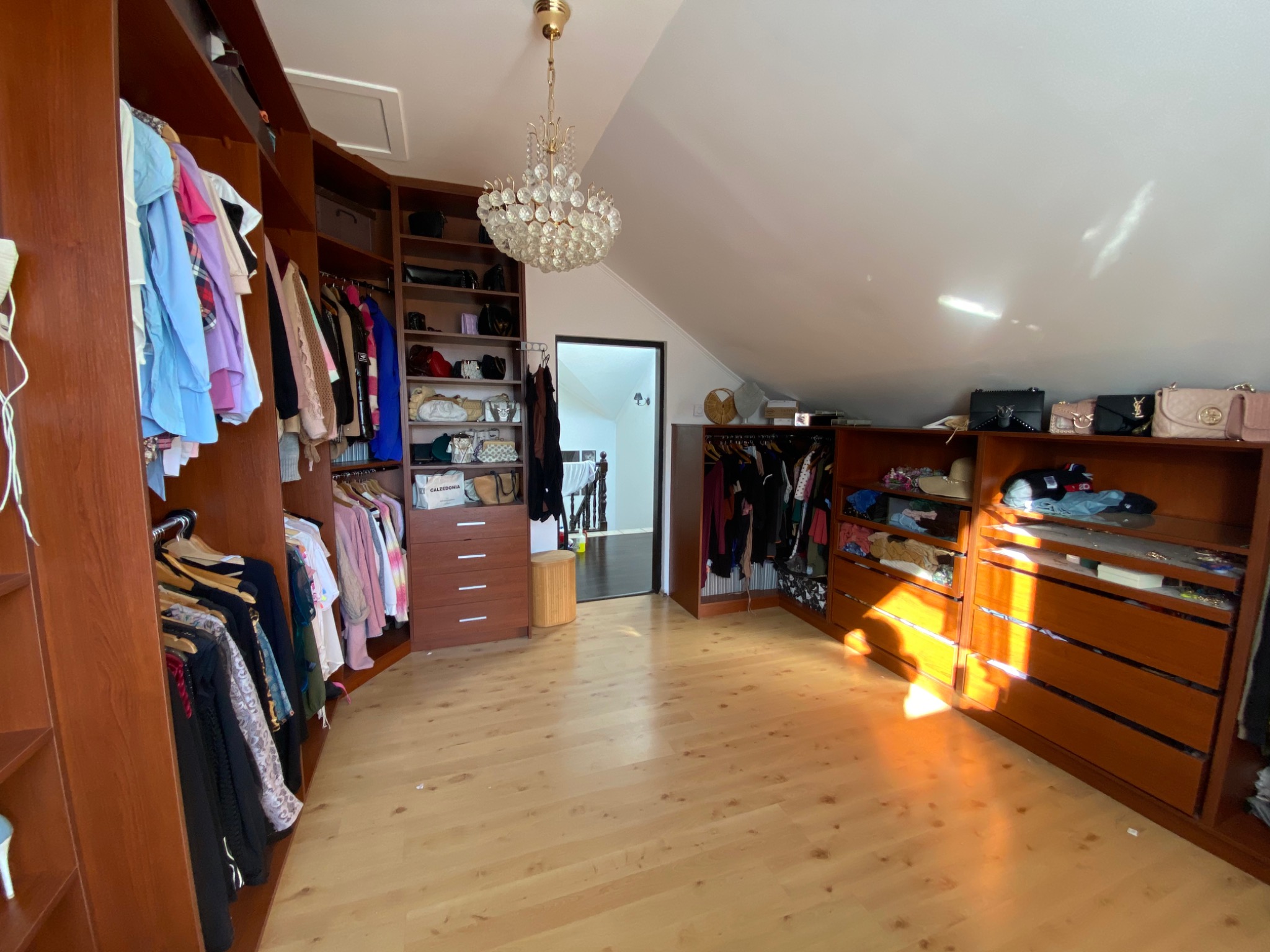 Przestronna garderoba na poddaszu z drewnianymi szafami, ubraniami na wieszakach, torebkami na półkach i kryształowym żyrandolem.