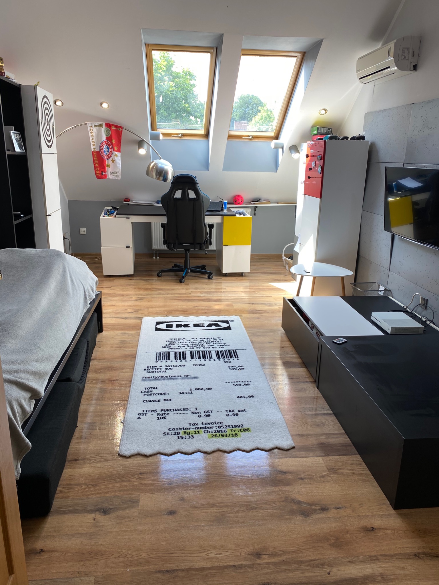 Pokój na poddaszu z drewnianą podłogą i dywanem w kształcie paragonu IKEA, widoczne okna dachowe, biurko z krzesłem gamingowym, łóżko i szafka RTV, na ścianie flaga Bayernu Monachium.
