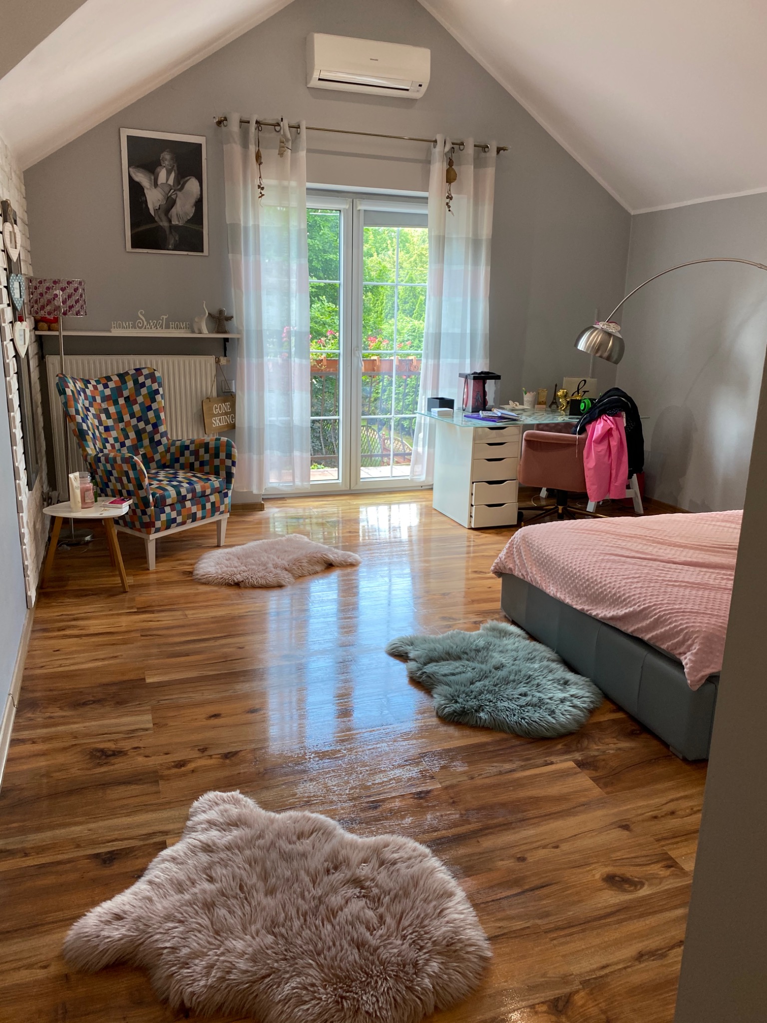 Jasna sypialnia na poddaszu z drewnianą podłogą, puszystymi dywanikami, łóżkiem, biurkiem i krzesłem, w tle okno balkonowe z widokiem na zieleń.