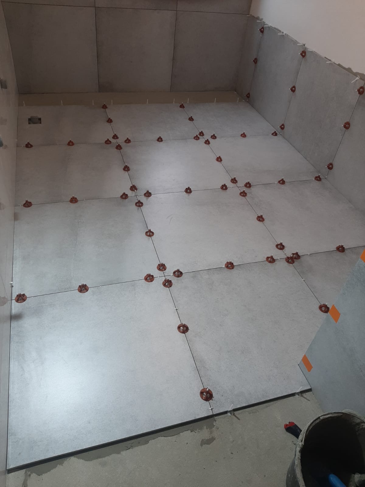 Szare płytki podłogowe w trakcie układania z użyciem systemu poziomowania płytek, widoczny fragment ściany z przyklejoną płytką z pomarańczowymi paskami taśmy.