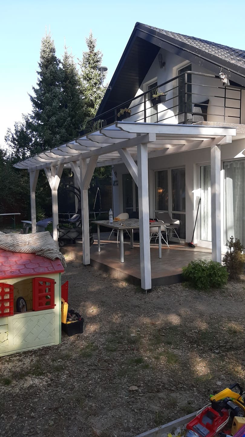 Biała pergola z przezroczystym zadaszeniem przylegająca do domu z balkonem, widoczny stół z krzesłami na tarasie, zabawki dziecięce na pierwszym planie, drzewa iglaste w tle.
