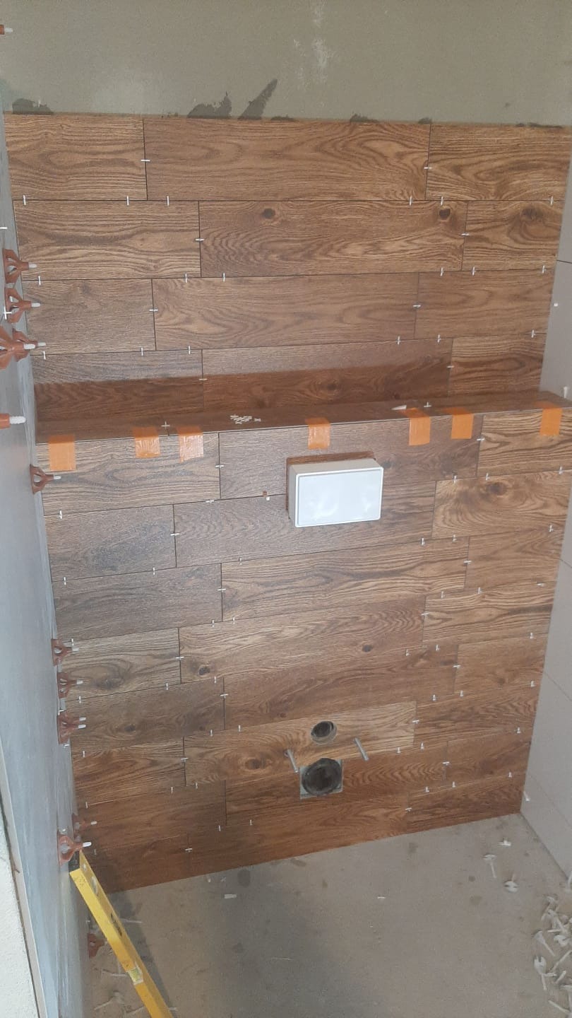 Ściana łazienki wyłożona brązowymi płytkami imitującymi drewno, z widocznymi krzyżykami dystansowymi, puszką elektryczną i otworem na rurę odpływową, w trakcie montażu.