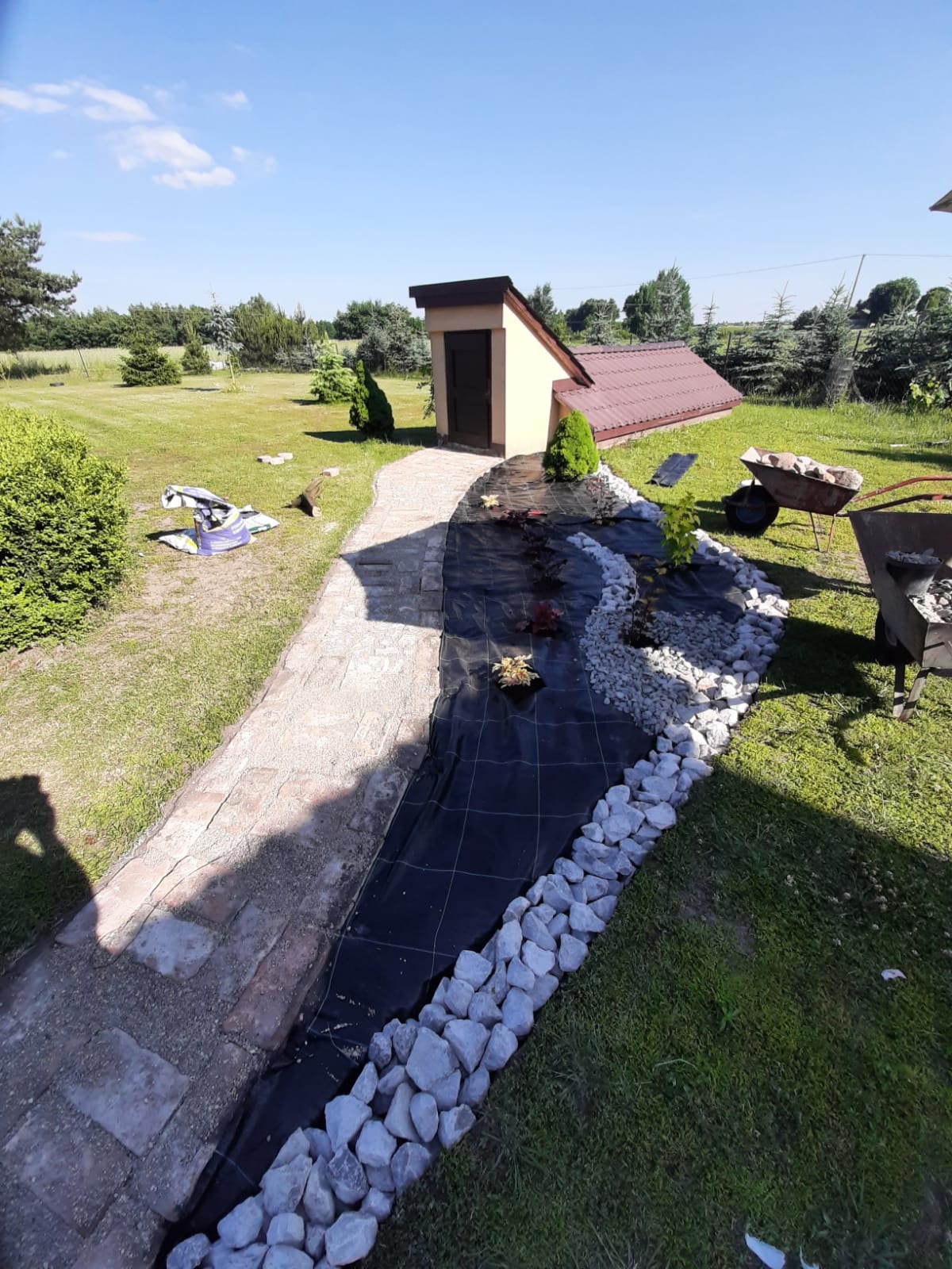 Tworzenie koryta strumienia w ogrodzie: czarna włóknina, białe kamienie, ścieżka z płyt, taczka z kamieniami, mały budynek gospodarczy w tle.