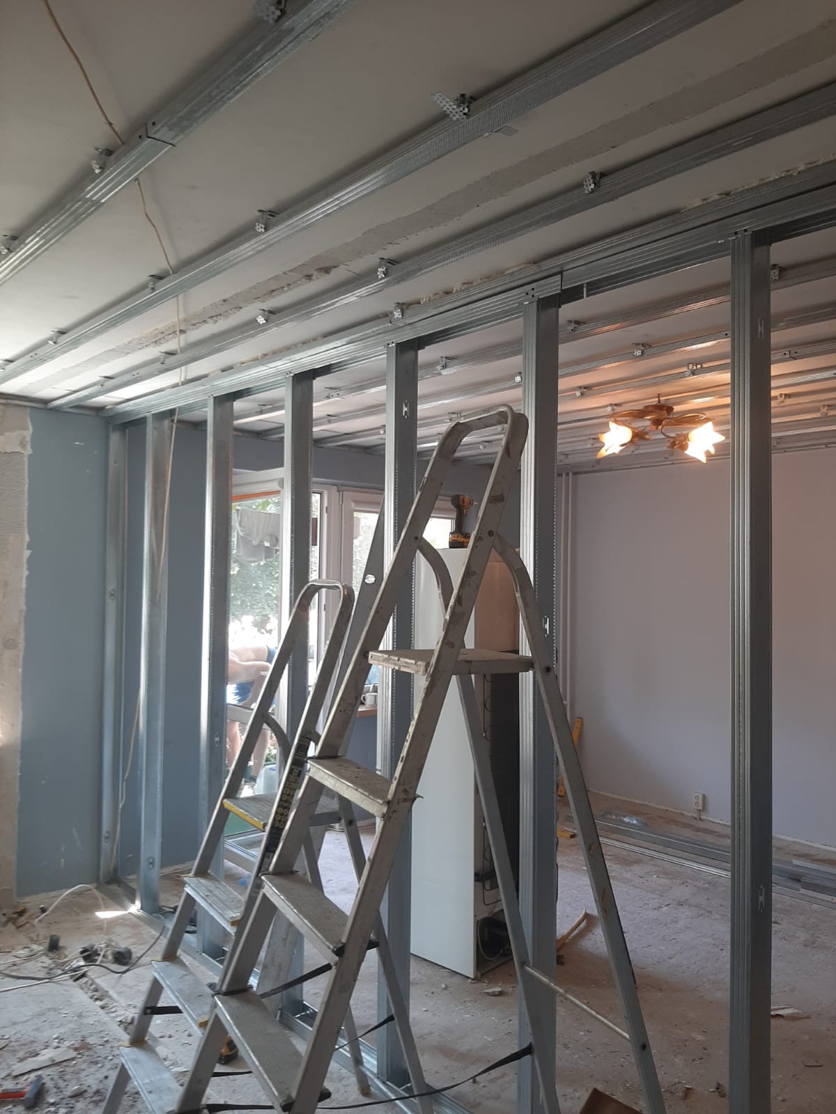 Montaż metalowej konstrukcji pod płytę karton gips na suficie i ścianach, widoczna drabina aluminiowa, w tle okno i lampa sufitowa.