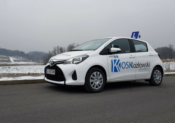 Biała Toyota Yaris z niebieskim logo i napisem 'KOS Kozłowski prawo jazdy kat. B' na boku, z literą 'L' na dachu, zaparkowana na asfalcie z ośnieżonym terenem w tle.