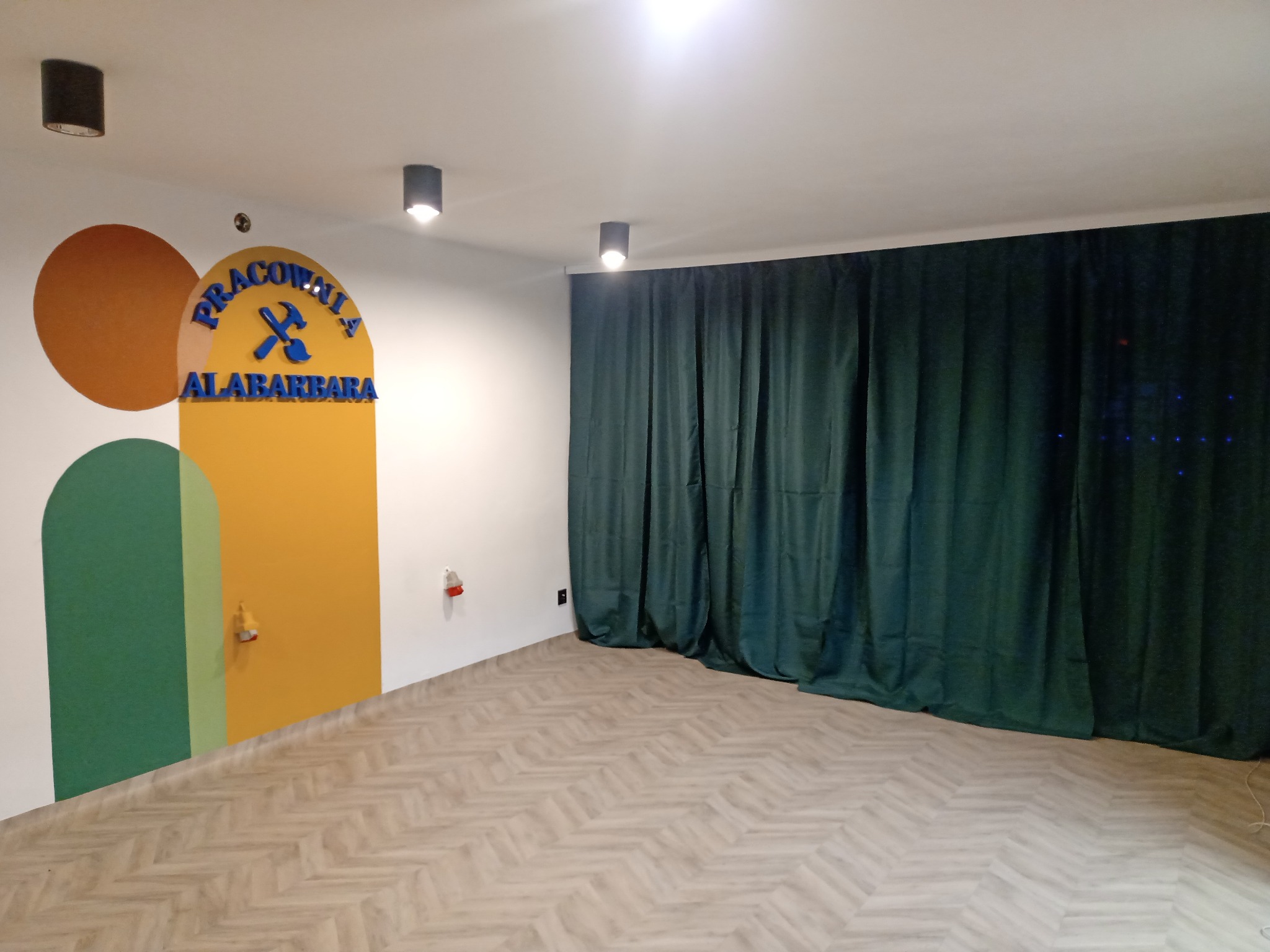 Pusta przestrzeń biurowa z parkietem w jodełkę, zasłonięta zielonymi zasłonami, z kolorową dekoracją ścienną i napisem 'Pracownia Alabarbara'.