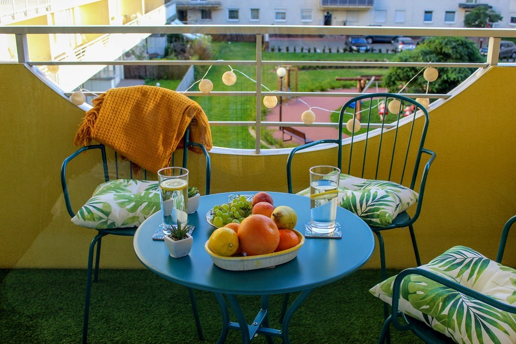 Turkusowy stolik z owocami i szklankami wody na balkonie z zieloną trawą, żółtą ścianą i dekoracyjnymi lampkami.