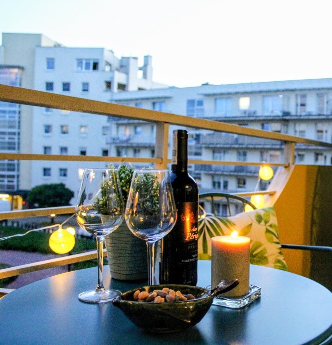 Stół na balkonie z dwoma kieliszkami, butelką wina, świecą i miseczką orzechów, oświetlony girlandą żarówkową, z widokiem na budynek mieszkalny w tle.