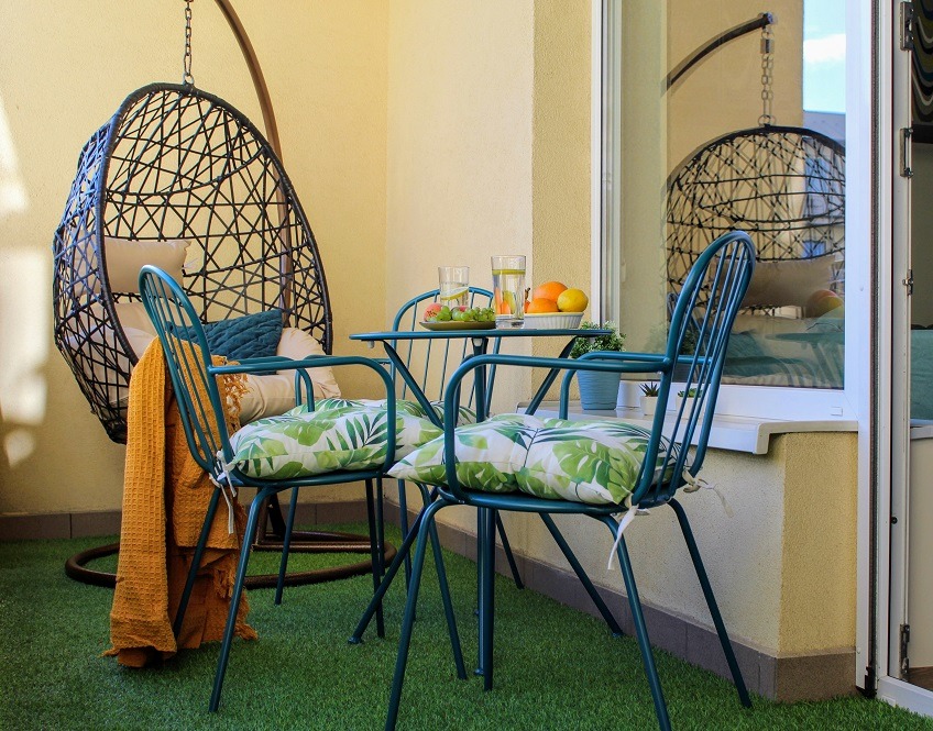 Balkon z zieloną sztuczną trawą, wiszącym fotelem kokon, metalowym stolikiem i krzesłami z poduszkami w liście, na stole owoce i woda w szklankach.